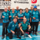Zdjęcie przedstawiające grupę seniorów w sportowych strojach pozujących w hali sportowej z kulami do gry w petanque, na tle banerów organizatorów wydarzenia.
