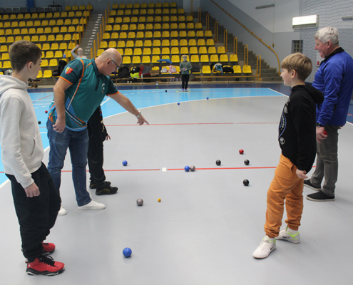 Galeria zdjęć z warsztatów soft petanque w hali sportowej, przedstawiająca dzieci i dorosłych podczas rzutów kolorowymi kulami oraz instruktażu prowadzonego przez opiekunów.