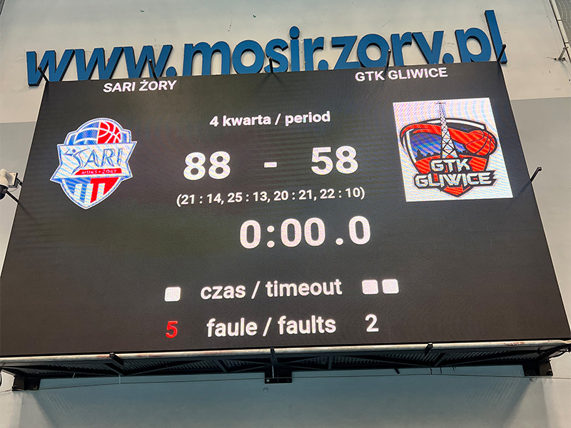 Zdjęcie tablicy wyników w hali sportowej pokazującej zakończony mecz koszykówki z wyraźnym wynikiem, logotypami drużyn z Żor i Gliwic oraz szczegółami czwartej kwarty.