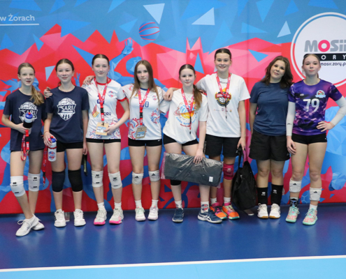 Galeria zdjęć z turnieju siatkówki dziewcząt w hali sportowej przedstawia drużyny pozujące na tle ścianki MOSiR, dynamiczne akcje przy siatce – bloki, ataki i obrony – oraz sportową rywalizację na tle żółtych trybun.