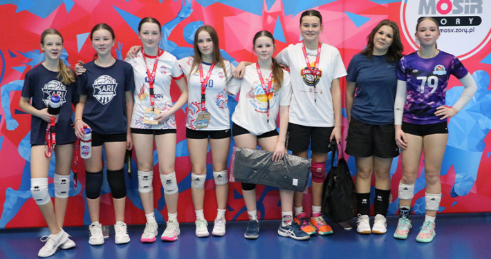 Galeria zdjęć z turnieju siatkówki dziewcząt w hali sportowej przedstawia drużyny pozujące na tle ścianki MOSiR, dynamiczne akcje przy siatce – bloki, ataki i obrony – oraz sportową rywalizację na tle żółtych trybun.