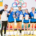 Zdjęcie grupowe zawodniczek i trenera podczas wydarzenia sportowego. Uczestniczki prezentują zdobyte medale, puchary oraz dyplomy.
