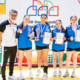 Zdjęcie grupowe zawodniczek i trenera podczas wydarzenia sportowego. Uczestniczki prezentują zdobyte medale, puchary oraz dyplomy.