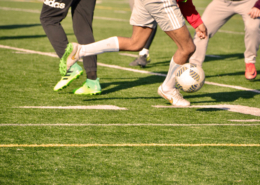 Na zdjęciu trzech piłkarzy grających w piłkę nożną na zielonej murawie, widoczne tylko nogi