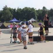 Na zdjęciu grupa dzieci z instruktorem na skate parku ćwicząca jazdę na hulajnodze