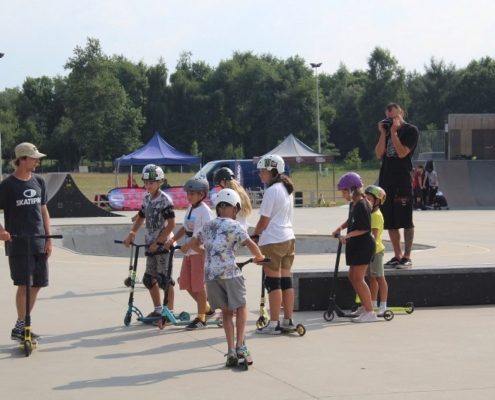 Na zdjęciu grupa dzieci z instruktorem na skate parku ćwicząca jazdę na hulajnodze