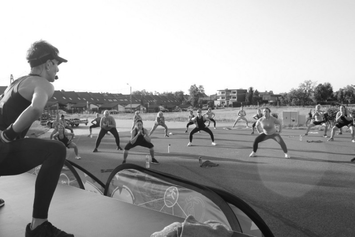 Na czarno-białym zdjęciu zajęcia fitness w parku cegielnia, w tle ćwiczący