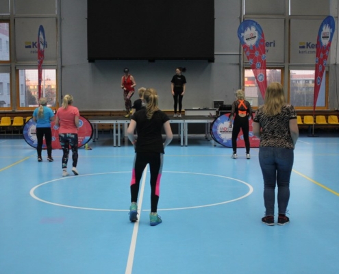 na zdjęciu uczestnicy zajęć na płycie hali sportowej ćwiczący przed sceną