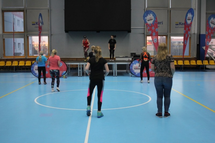 na zdjęciu uczestnicy zajęć na płycie hali sportowej ćwiczący przed sceną