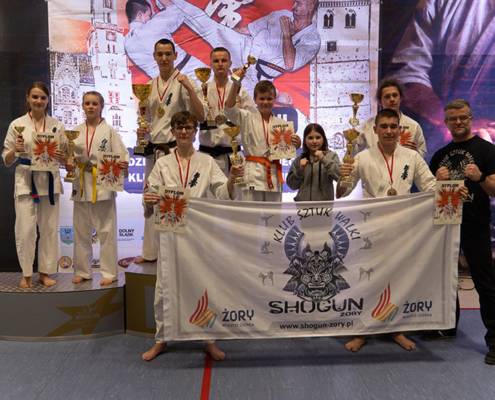 Na zdjęciu widać grupę młodych zawodników karate z Klubu Sztuk Walki Shogun Żory, stojących na podium z pucharami, medalami i dyplomami.