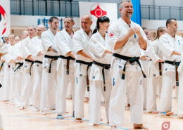 Zdjęcia przedstawiają treningi i pokazy karate, zarówno w hali sportowej, jak i na świeżym powietrzu, z udziałem wielu osób w białych strojach, wykonujących ćwiczenia, walki sparingowe oraz pozujących do zdjęć grupowych.