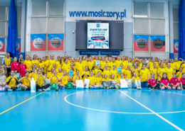 galeria zdjęć przedstawiającej relację z wydarzenia sportowego w hali MOSiR, na którym widać dzieci i młodzież w żółtych koszulkach podczas występów, wręczania nagród i wspólnego ustawienia na płycie boiska.