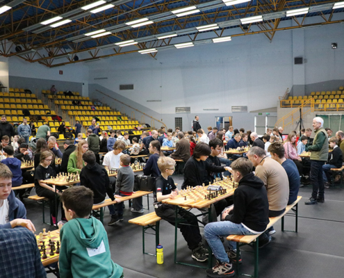 Galeria zdjęć z Otwartego Turnieju Szachowego w hali sportowej – rozgrywki dzieci i młodzieży przy szachownicach, emocjonujące pojedynki w różnych kategoriach wiekowych, ceremonia dekoracji zwycięzców oraz organizacja wydarzenia przy stoliku sędziowskim.