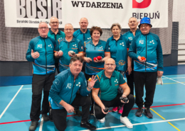 Zdjęcie przedstawiające grupę seniorów w sportowych strojach pozujących w hali sportowej z kulami do gry w petanque, na tle banerów organizatorów wydarzenia.