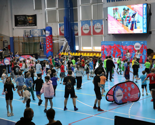 Galeria zdjęć z wydarzenia sportowo-rekreacyjnego w hali, przedstawiająca dzieci uczestniczące w konkurencjach sprawnościowych, zabawach na dmuchańcach oraz animacjach prowadzonych na płycie boiska.