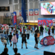 Galeria zdjęć z wydarzenia sportowo-rekreacyjnego w hali, przedstawiająca dzieci uczestniczące w konkurencjach sprawnościowych, zabawach na dmuchańcach oraz animacjach prowadzonych na płycie boiska.