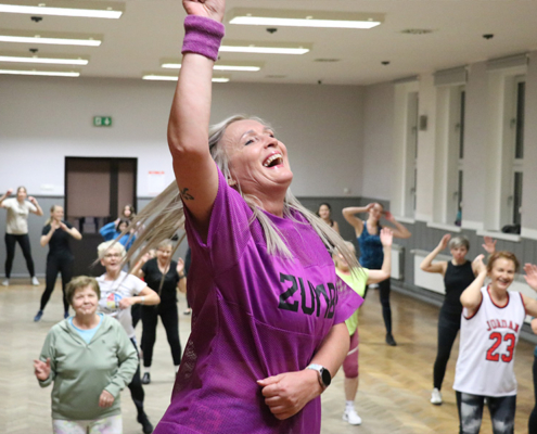 Galeria zdjęć z zajęć taneczno-fitnessowych, przedstawiająca instruktorkę prowadzącą energetyczny trening w sali oraz dzieci i dorosłych ćwiczących wspólnie choreografię.