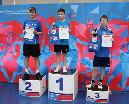 Galeria zdjęć z turnieju tenisa stołowego w hali sportowej – mecze dzieci i młodzieży przy stołach, dynamiczne wymiany, szerokie ujęcia rozgrywek oraz ceremonia dekoracji zwycięzców na podium.