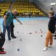 Galeria zdjęć z warsztatów soft petanque w hali sportowej, przedstawiająca dzieci i dorosłych podczas rzutów kolorowymi kulami oraz instruktażu prowadzonego przez opiekunów.