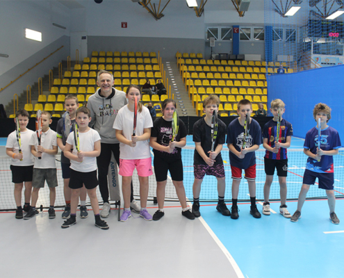 Galeria zdjęć z turnieju tenisa stołowego w hali sportowej, przedstawiająca dzieci ustawione do wręczenia nagród oraz uczestników rozgrywających mecze pod okiem trenera.