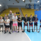 Galeria zdjęć z turnieju tenisa stołowego w hali sportowej, przedstawiająca dzieci ustawione do wręczenia nagród oraz uczestników rozgrywających mecze pod okiem trenera.