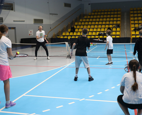 Zdjęcie przedstawiające dzieci uczestniczące w zajęciach tenisowych w hali sportowej, ćwiczące odbicia piłek pod okiem trenera na wydzielonych kortach z niską siatką.