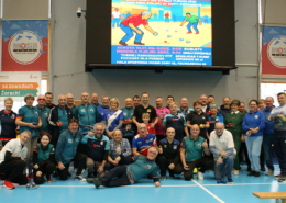 Galeria zdjęć z turnieju sportowego seniorów w hali MOSiR przedstawia uczestników w sportowych koszulkach podczas wręczania pucharów i dyplomów, wspólne fotografie zwycięzców oraz momenty dekoracji na tle siatki i hali sportowej.