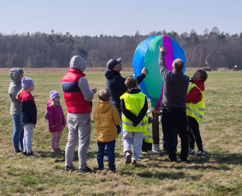 Zestaw zdjęć z wydarzenia plenerowego na lotnisku, przedstawiających dzieci i dorosłych wypuszczających kolorowe balony (m.in. czerwone, żółte, niebieskie) na tle otwartego pola i hangaru, a także grupowe ujęcia z dyplomami w jasnej, słonecznej scenerii.