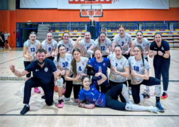 Zdjęcie drużyny siatkarskiej kobiet w hali sportowej, pozującej wspólnie z trenerem w dynamicznych, radosnych pozach po meczu.