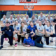 Zdjęcie drużyny siatkarskiej kobiet w hali sportowej, pozującej wspólnie z trenerem w dynamicznych, radosnych pozach po meczu.
