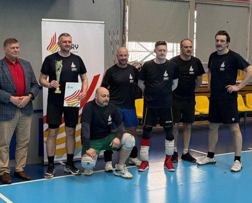 Zdjęcie drużyny uczestników w hali sportowej podczas ceremonii zakończenia rozgrywek, z zawodnikiem trzymającym puchar i dyplom.