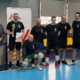 Zdjęcie drużyny uczestników w hali sportowej podczas ceremonii zakończenia rozgrywek, z zawodnikiem trzymającym puchar i dyplom.