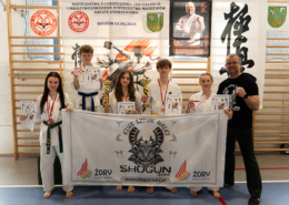 Galeria przedstawia zawody sztuk walki w hali sportowej – ujęcia z walk na macie w kaskach i ochraniaczach, momenty wręczenia nagród na podium oraz wspólne zdjęcia uczestników w kimonach.