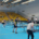 Galeria zdjęć przedstawiająca rozgrywki sportowe w hali. Widoczni są uczestnicy grający przy stołach, zawodnicy ustawieni na boisku, wręczenie nagród na podium oraz rywalizacja przy planszach.
