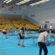 Galeria zdjęć przedstawiająca rozgrywki sportowe w hali. Widoczni są uczestnicy grający przy stołach, zawodnicy ustawieni na boisku, wręczenie nagród na podium oraz rywalizacja przy planszach.