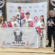 Galeria zdjęć z wydarzenia sportowego odbywającego się w hali. Widoczni są uczestnicy w strojach do sztuk walki, pokazy, wspólne ustawienia zawodników oraz wręczanie nagród.