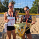 Dwie młode siatkarki plażowe w czarnych strojach sportowych pozują na boisku z pucharem, medalami, dyplomem i statuetką za zdobyte pierwsze miejsce. Na pierwszym planie znajduje się duży złoty balon w kształcie pucharu z cyfrą „1”, a w tle widoczne jest boisko do siatkówki plażowej z siatką.